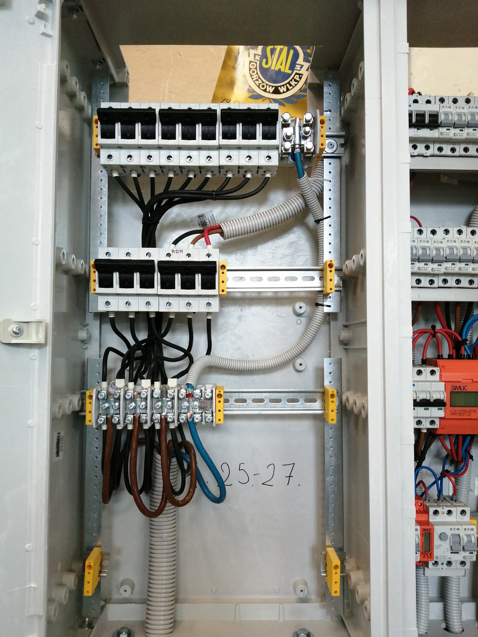Rozdzielnica elektryczna w trakcie montażu, widoczne przewody, bezpieczniki i listwy zaciskowe. Elementy instalacji wewnątrz obudowy, z napisem '25-27' na ściance.