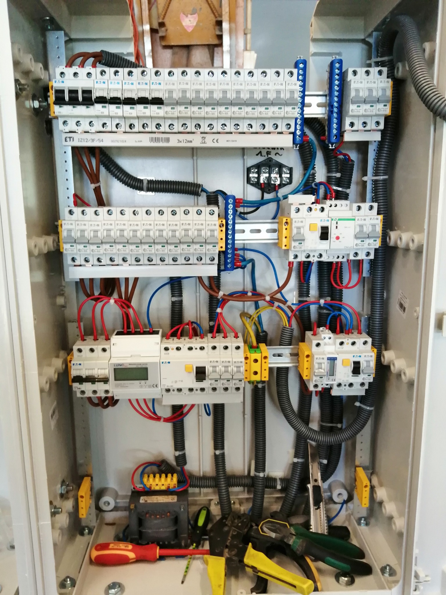 Rozdzielnica elektryczna w obudowie, widoczne zabezpieczenia, przewody, listwy zaciskowe i narzędzia monterskie na dnie szafy. Instalacja wykonana schludnie, przewody uporządkowane.