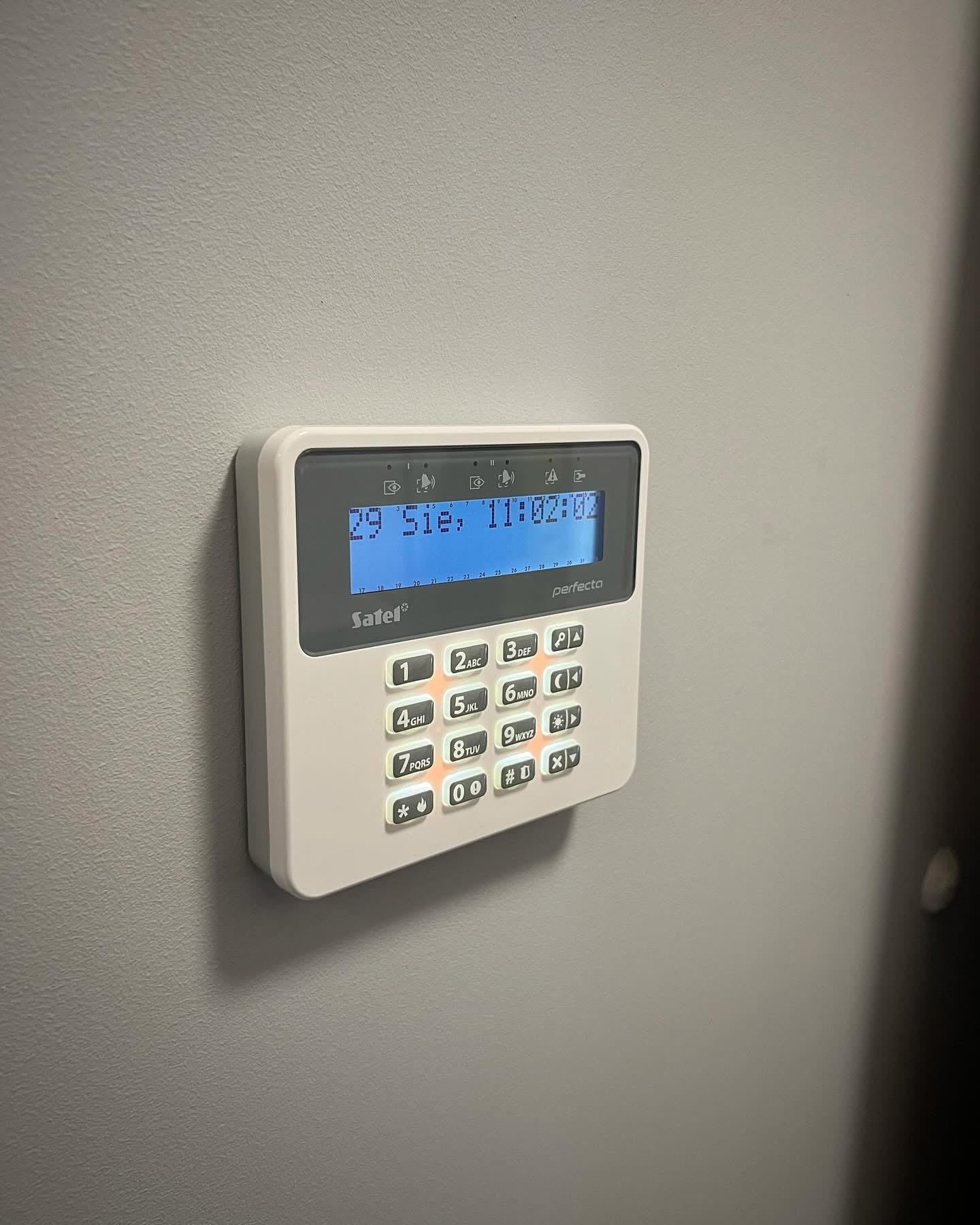 Panel sterowania alarmem Satel Perfecta zamontowany na szarej ścianie, z widocznym wyświetlaczem i klawiaturą numeryczną. Data i godzina na ekranie: 29 Sie, 11:02:02.