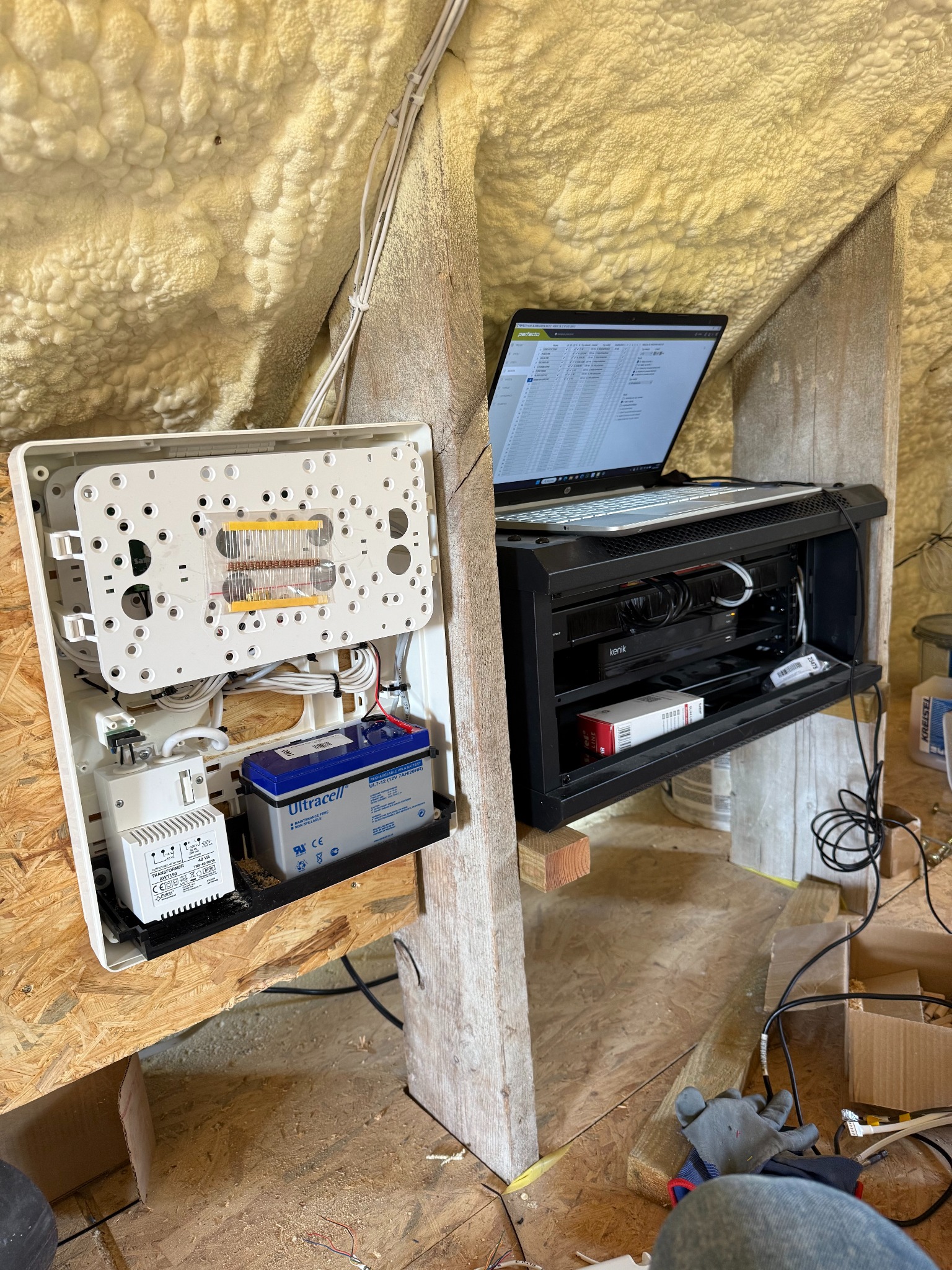 Centrala alarmowa z akumulatorem, laptop i sprzęt sieciowy w szafce rack na poddaszu z izolacją piankową. Widoczne kable i narzędzia.