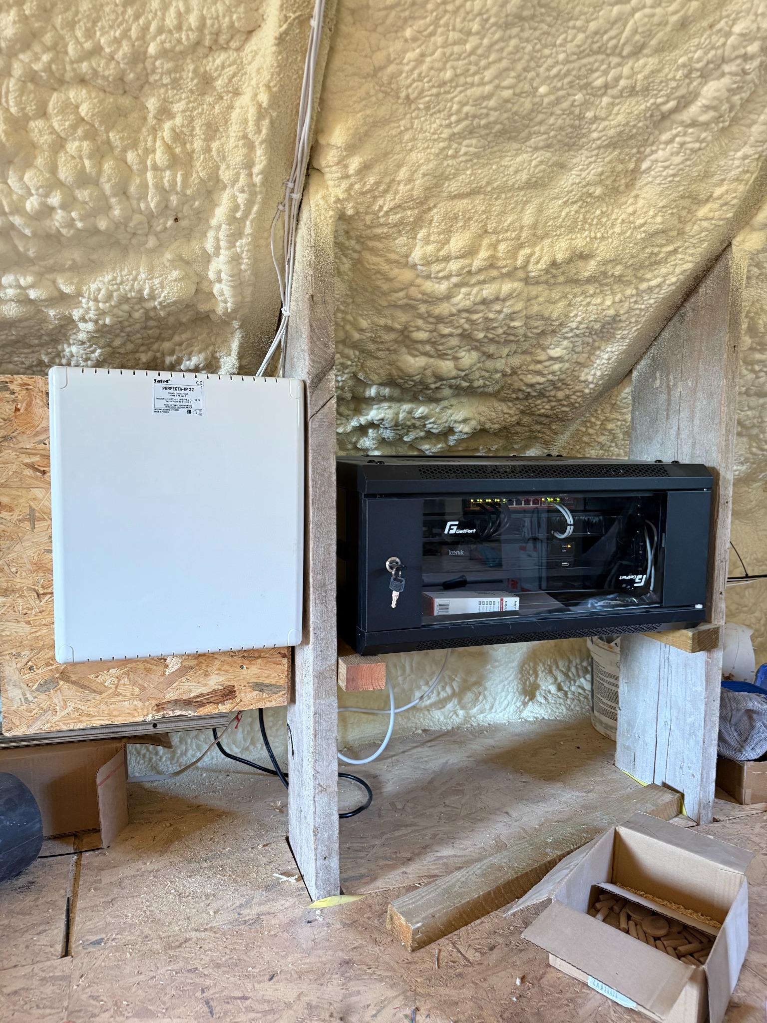 Zamontowana szafa rack z osprzętem telekomunikacyjnym i centrala alarmowa na drewnianej konstrukcji w pomieszczeniu z izolacją piankową.