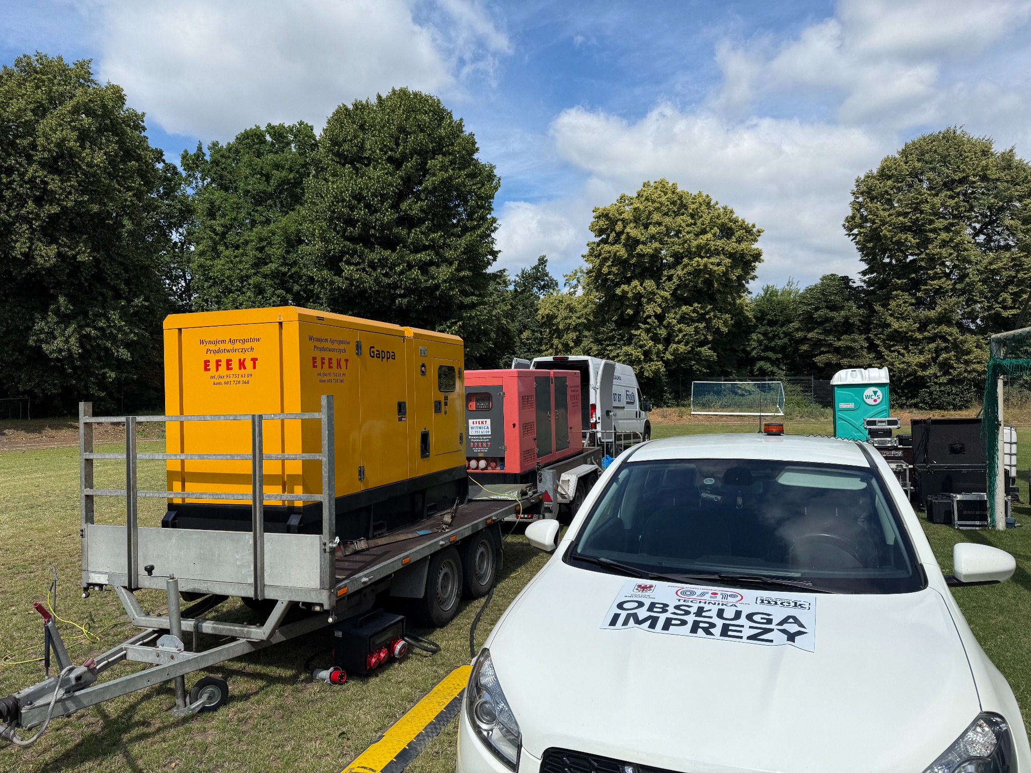 Agregaty prądotwórcze na przyczepie, z żółtą obudową, na trawiastym terenie, z samochodem 'Obsługa Imprez' na pierwszym planie. Widoczne drzewa i niebo.