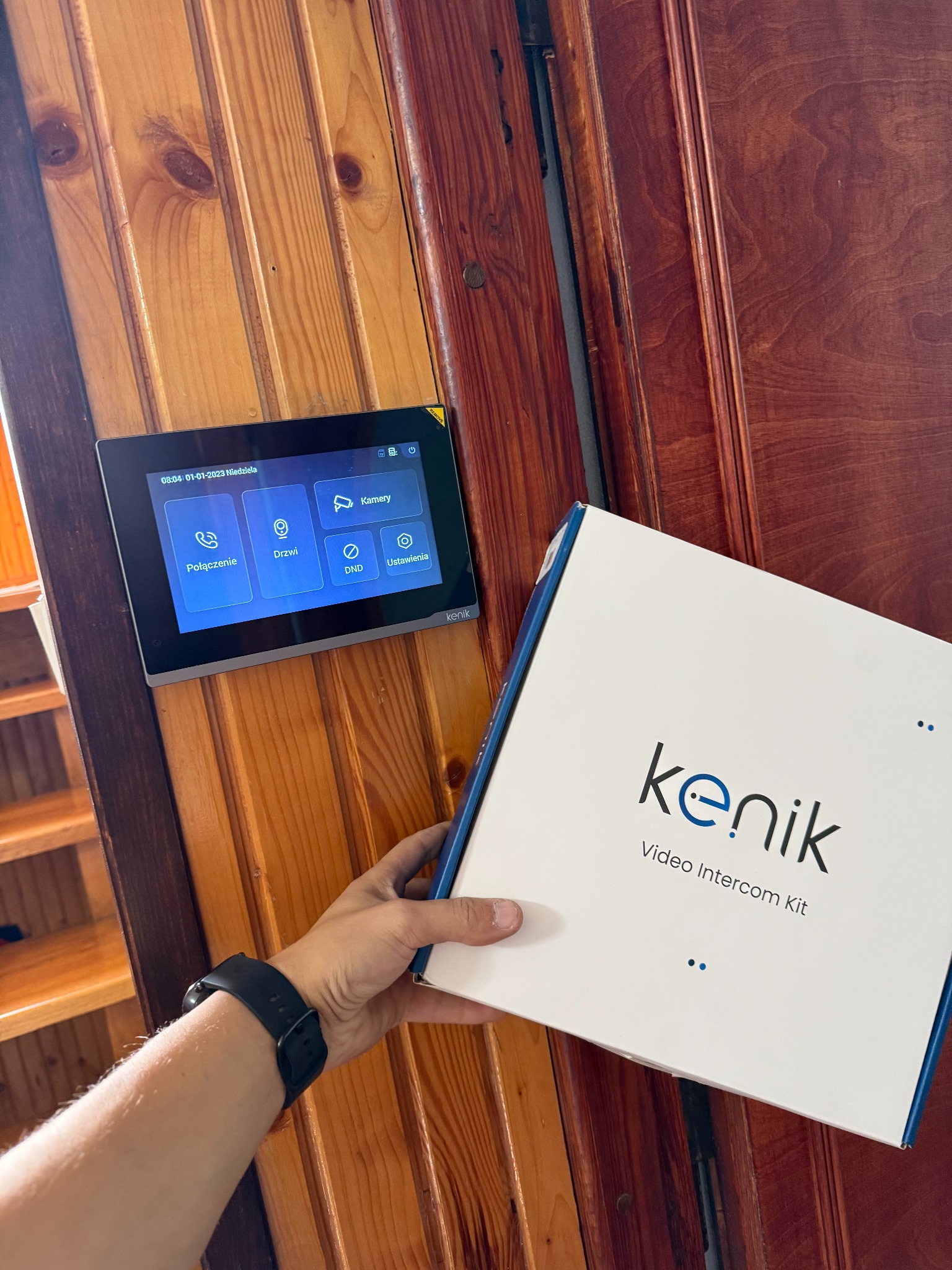 Zamontowany panel wideodomofonu Kenik na drewnianej ścianie, obok pudełko z zestawem. Widoczny fragment ręki z zegarkiem. Ustawienia: połączenie, drzwi, kamery.
