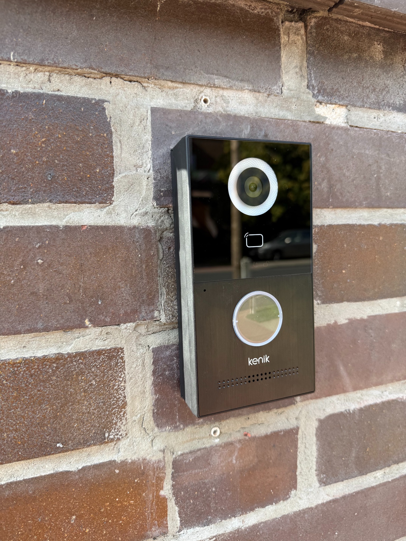 Czarny wideodomofon Kenik zamontowany na ceglanej ścianie zewnętrznej budynku. Widoczny obiektyw kamery, czytnik kart i przycisk dzwonka. Tło rozmyte, zarys samochodu.