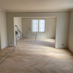 Wk Home - Jasny parkiet ułożony w jodełkę w przestronnym, odświeżonym wnętrzu. Widoczne kable elektryczne zwisające z sufitu, okno zabezpieczone folią malarską. Świeżo wykończone ściany.