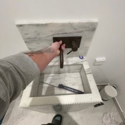 Wk Home - Marmurowa umywalka z brązową armaturą, montowana przez glazurnika. Widoczne narzędzia i fragment podłogi z płytek. Ujęcie z góry, podkreślające detale wykończenia.