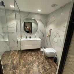 Wk Home - Nowoczesna łazienka z marmurowymi płytkami, brązową podłogą i okrągłym lustrem. Kabina prysznicowa ze szklanymi ścianami, biała umywalka z szafką i wisząca toaleta.