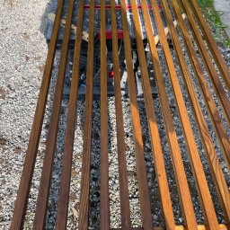 Wk Home - Drewniane, pomalowane listwy ułożone równolegle na czarnym stole roboczym, na tle żwirowego podłoża i trawy. Widoczne słoje drewna i świeży lakier.