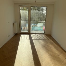 Wk Home - Jasne panele ułożone w jodełkę w pustym pokoju z balkonem. Świeże, białe ściany i grzejnik. Promienie słoneczne podkreślają wzór podłogi.