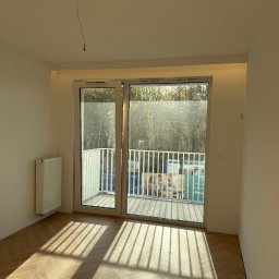 Wk Home - Jasne, puste wnętrze z parkietem ułożonym w jodełkę, balkonem z widokiem na zieleń i grzejnikiem. Promienie słoneczne wpadają przez okno.