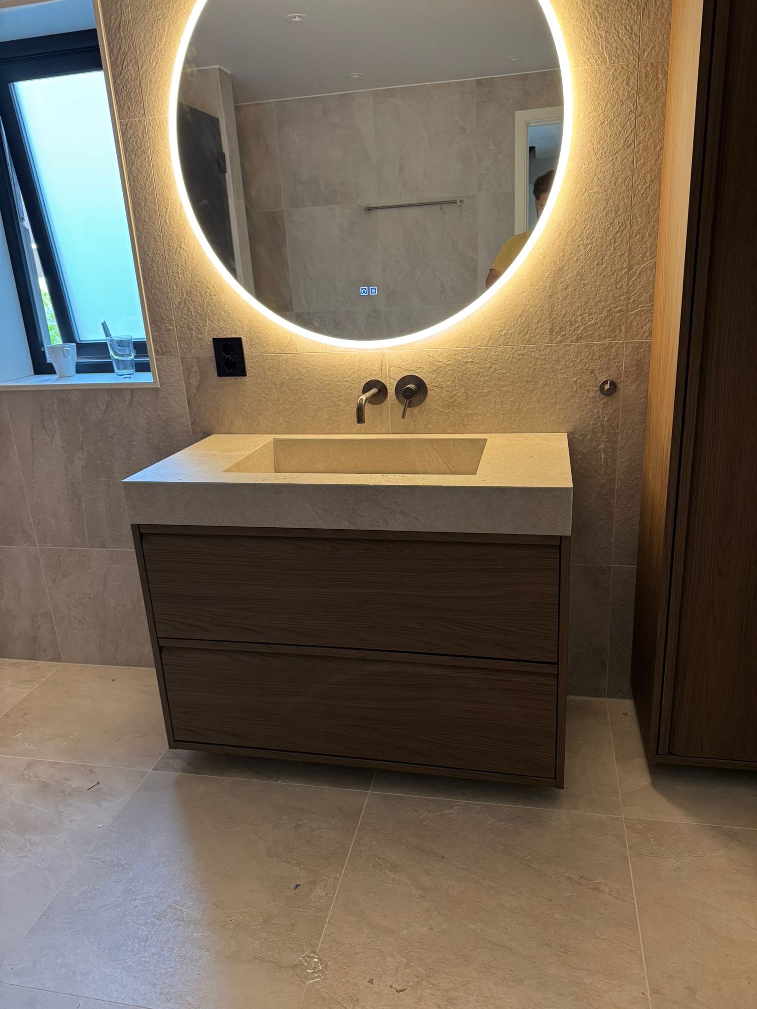 Nowoczesna łazienka: drewniana szafka z kamiennym blatem i okrągłym lustrem z podświetleniem LED. Beżowe płytki na ścianach i podłodze. Minimalistyczny design.