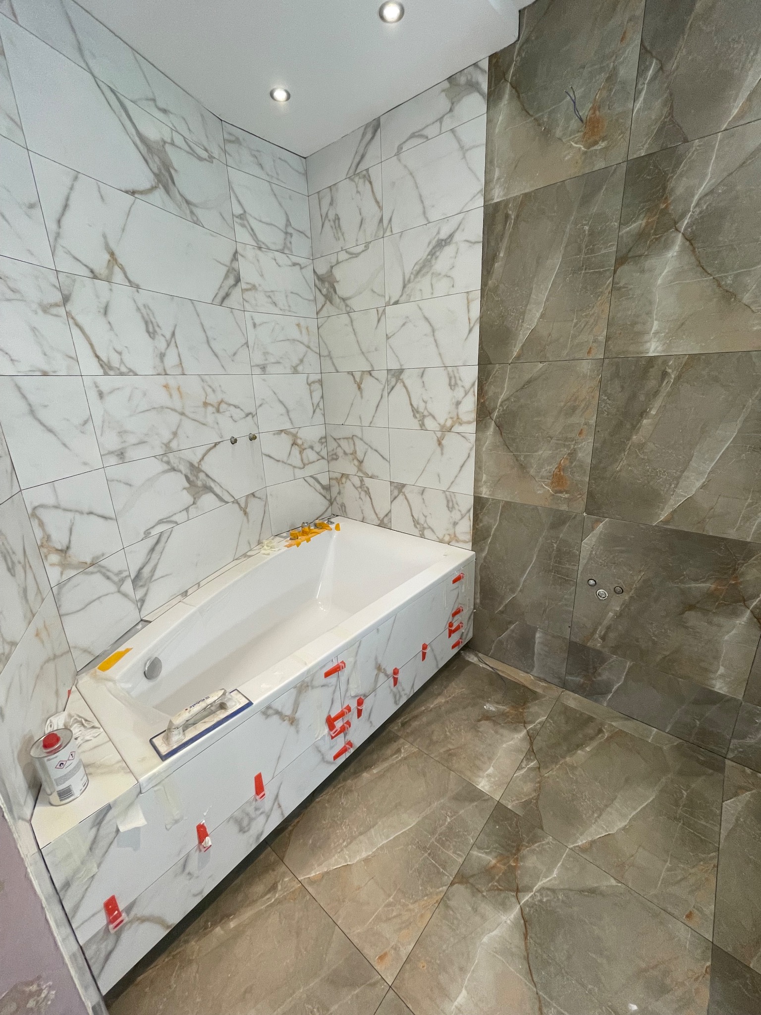 Biała wanna obudowana marmurowymi płytkami w łazience, część ścian wykończona szarymi płytkami z marmurowym wzorem. Widoczne narzędzia glazurnicze i krzyżyki dystansowe.