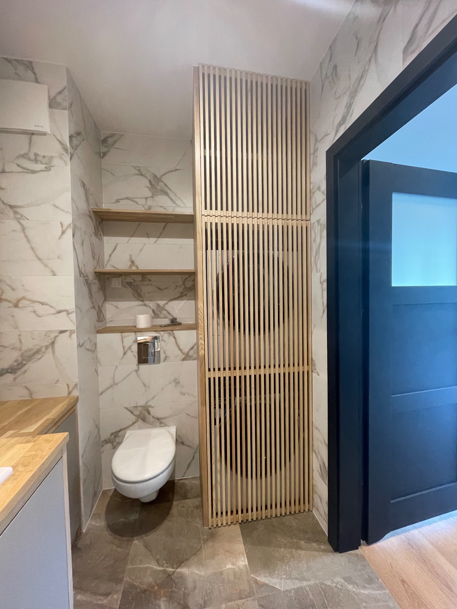 Nowoczesna łazienka z marmurowymi płytkami, drewnianymi półkami i ażurową ścianką działową z jasnego drewna, oddzielającą toaletę od reszty pomieszczenia. Ciemne drzwi.