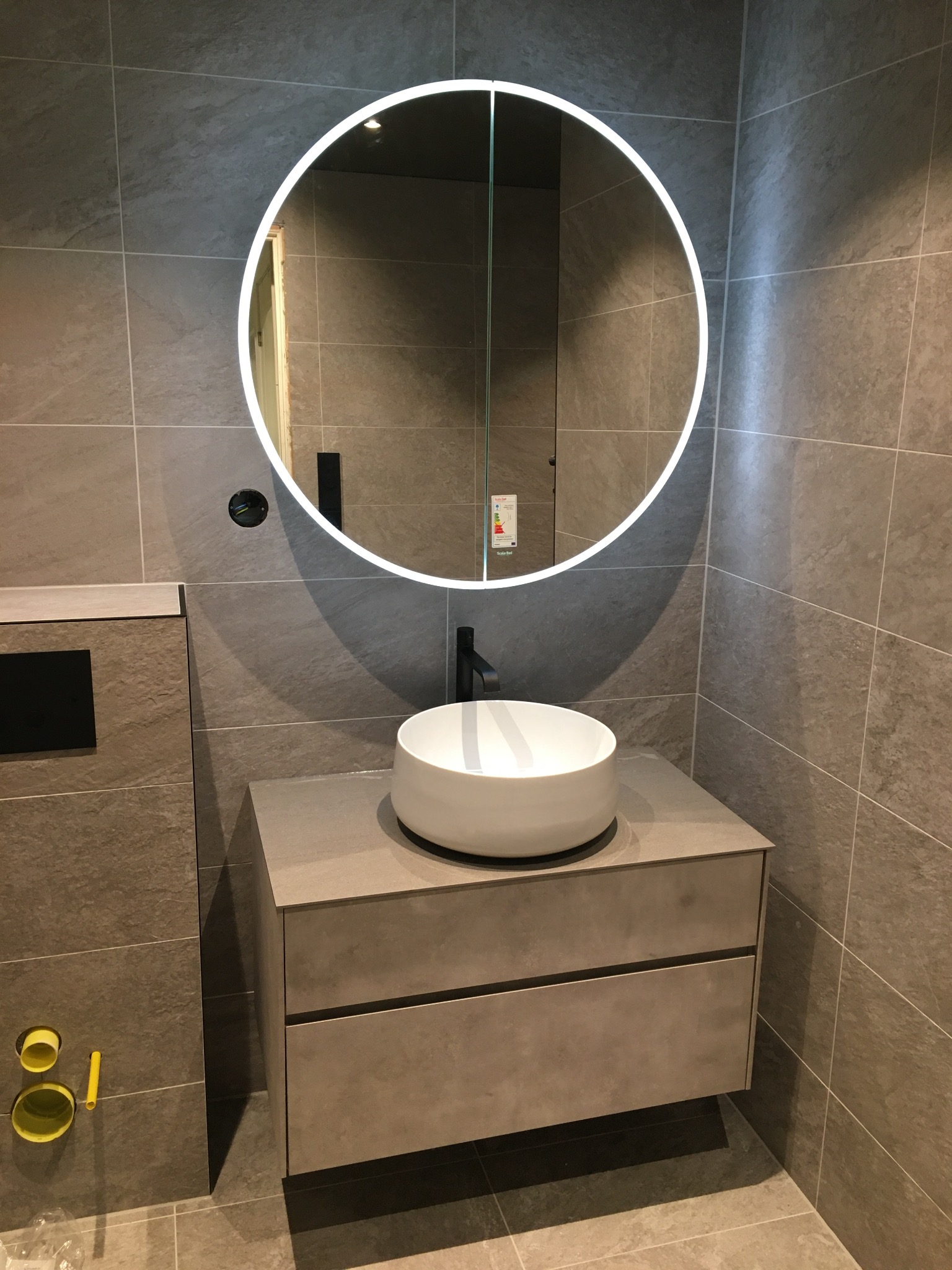 Nowoczesna łazienka w Gdyni: szare płytki, okrągłe lustro z podświetleniem LED, biała umywalka nablatowa na szafce z szufladami. Minimalistyczny design.