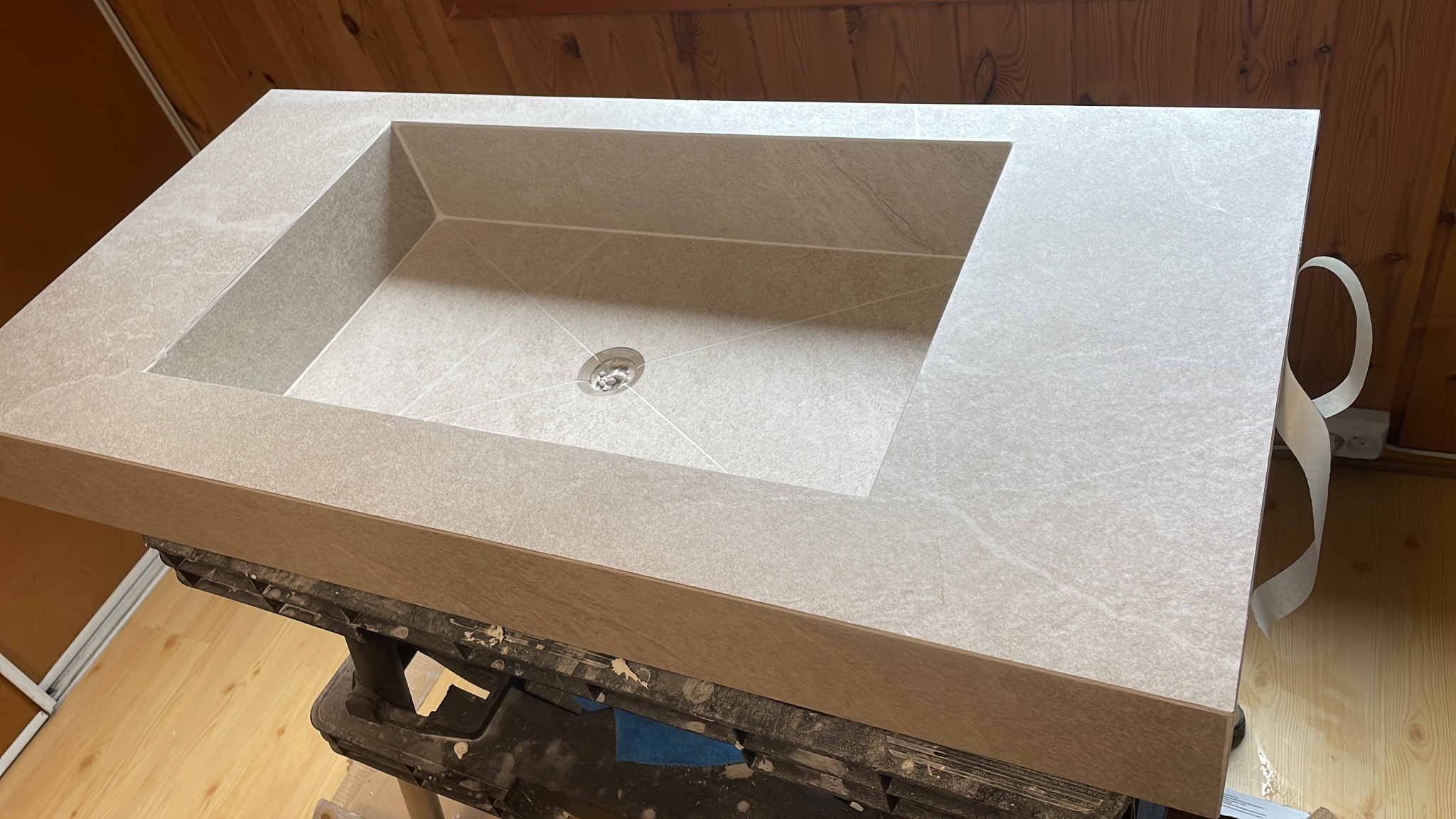 Beżowa umywalka z płytek, wbudowana w blat, z widocznym odpływem i fugami. Ustawiona na roboczym stole, gotowa do montażu w łazience. Minimalistyczny design.