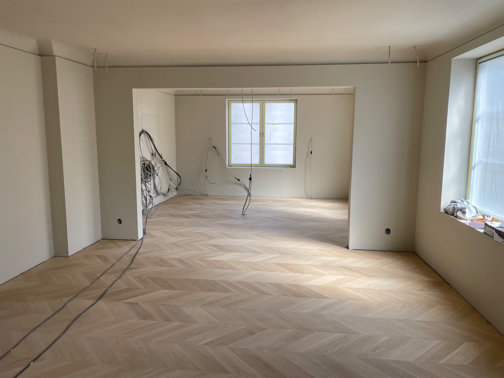 Jasny parkiet ułożony w jodełkę w przestronnym, odświeżonym wnętrzu. Widoczne kable elektryczne zwisające z sufitu, okno zabezpieczone folią malarską. Świeżo wykończone ściany.