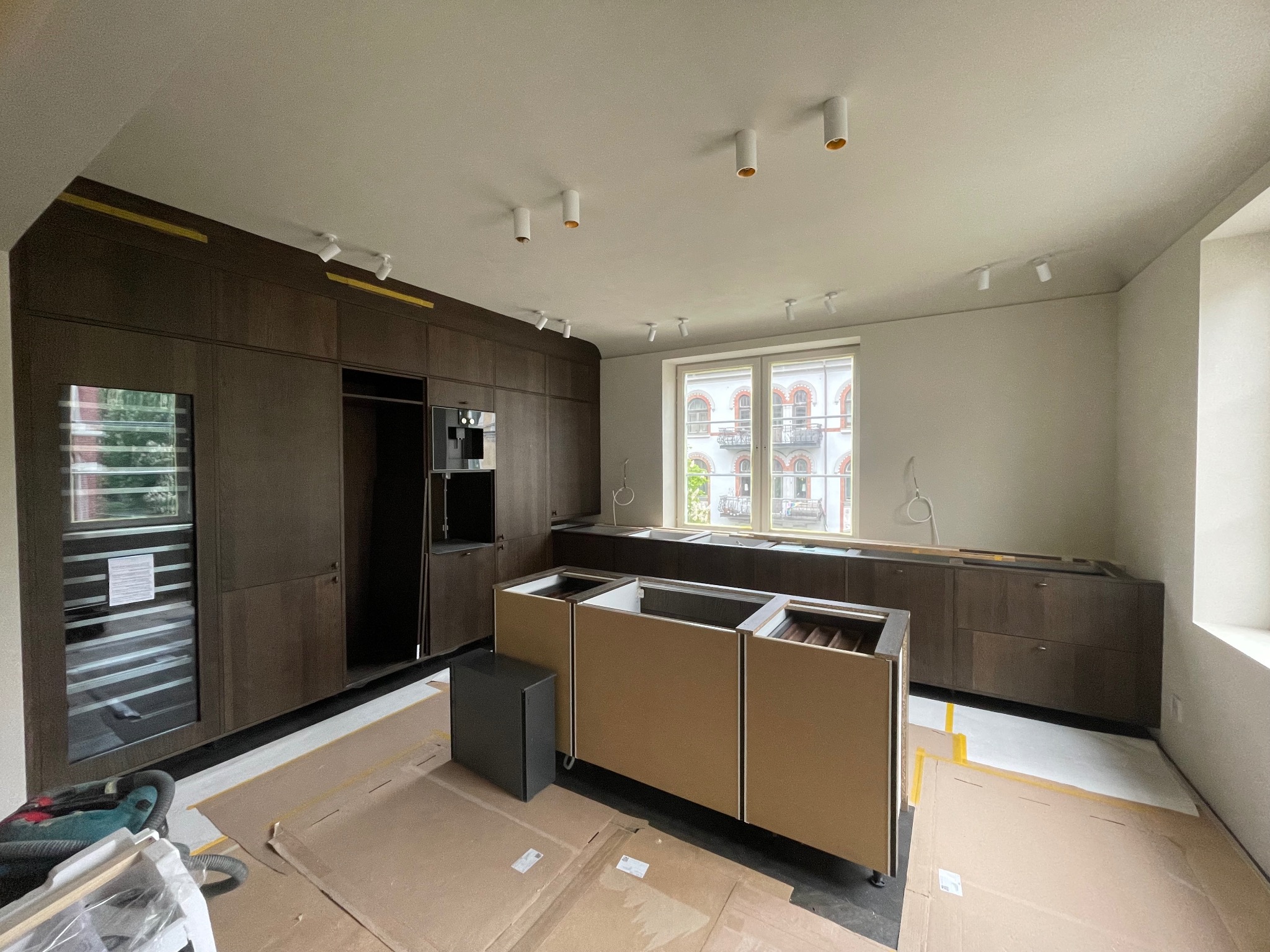 Częściowo zamontowana, nowoczesna kuchnia z ciemnymi szafkami i wyspą na kartonach ochronnych. Widoczne narzędzia i oświetlenie punktowe.