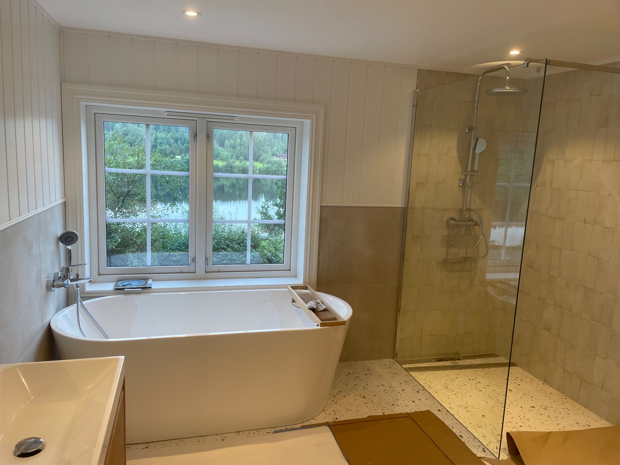 Jasna łazienka z wanną wolnostojącą i kabiną prysznicową typu walk-in, widok na jezioro przez okno. Minimalistyczny design, płytki w odcieniach beżu, armatura chromowana.