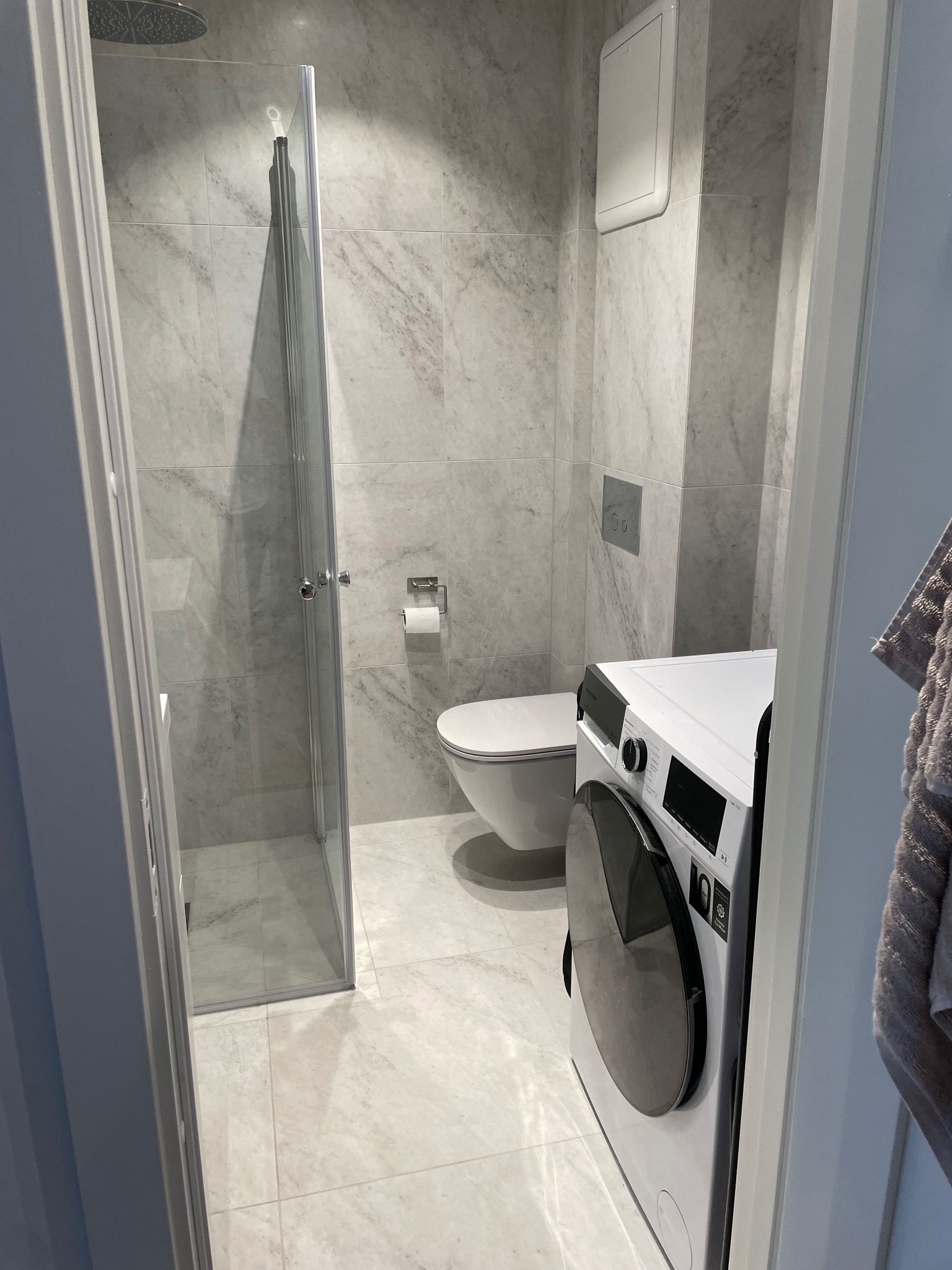 Jasna łazienka z kabiną prysznicową typu walk-in, toaletą i pralką. Ściany i podłoga wyłożone płytkami imitującymi marmur. Minimalistyczny design.