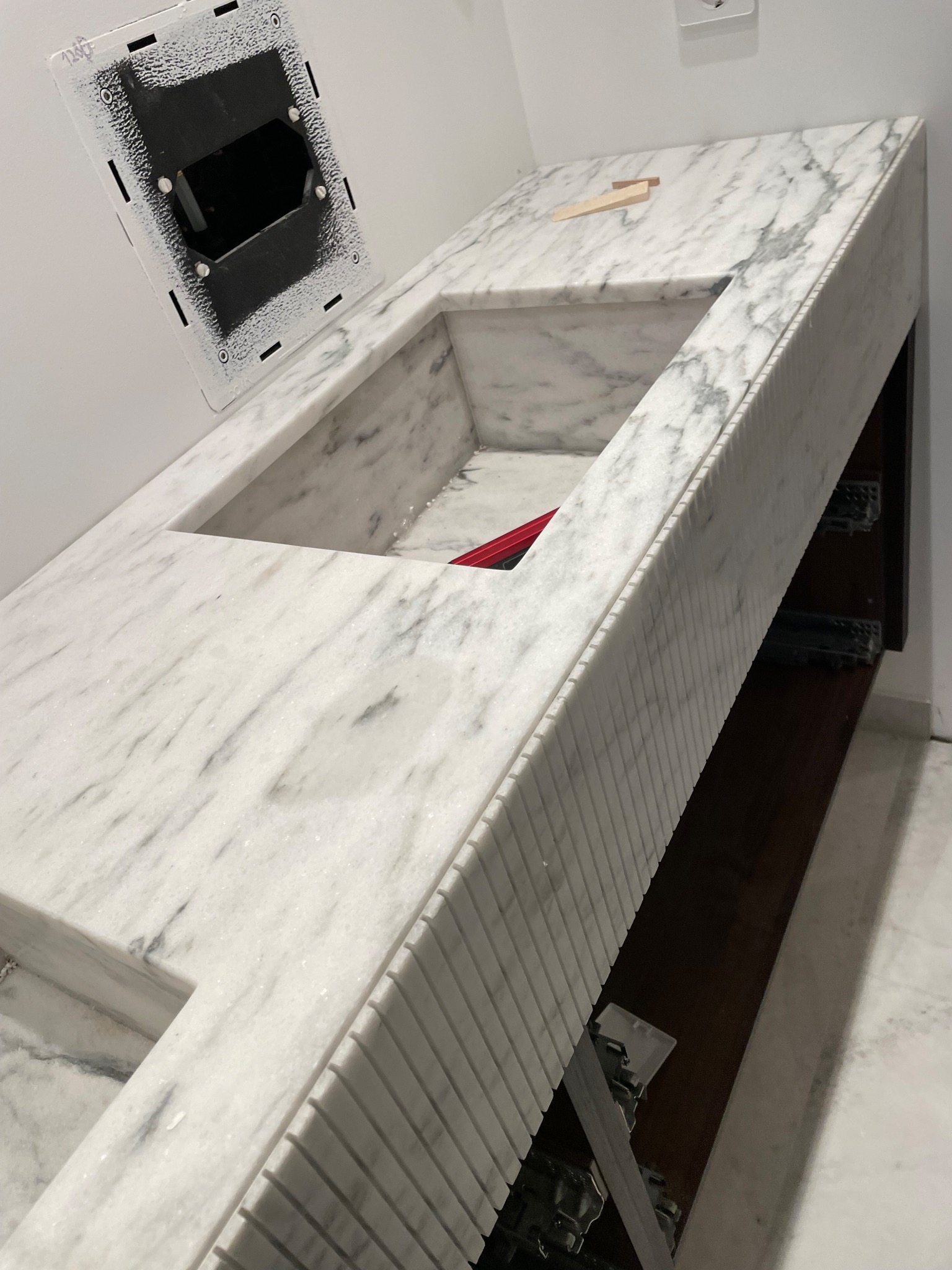 Marmurowa umywalka w łazience z dekoracyjnym frezowaniem na krawędziach, widoczna puszka elektryczna i drewniany klin. Ujęcie z góry, jasne oświetlenie.