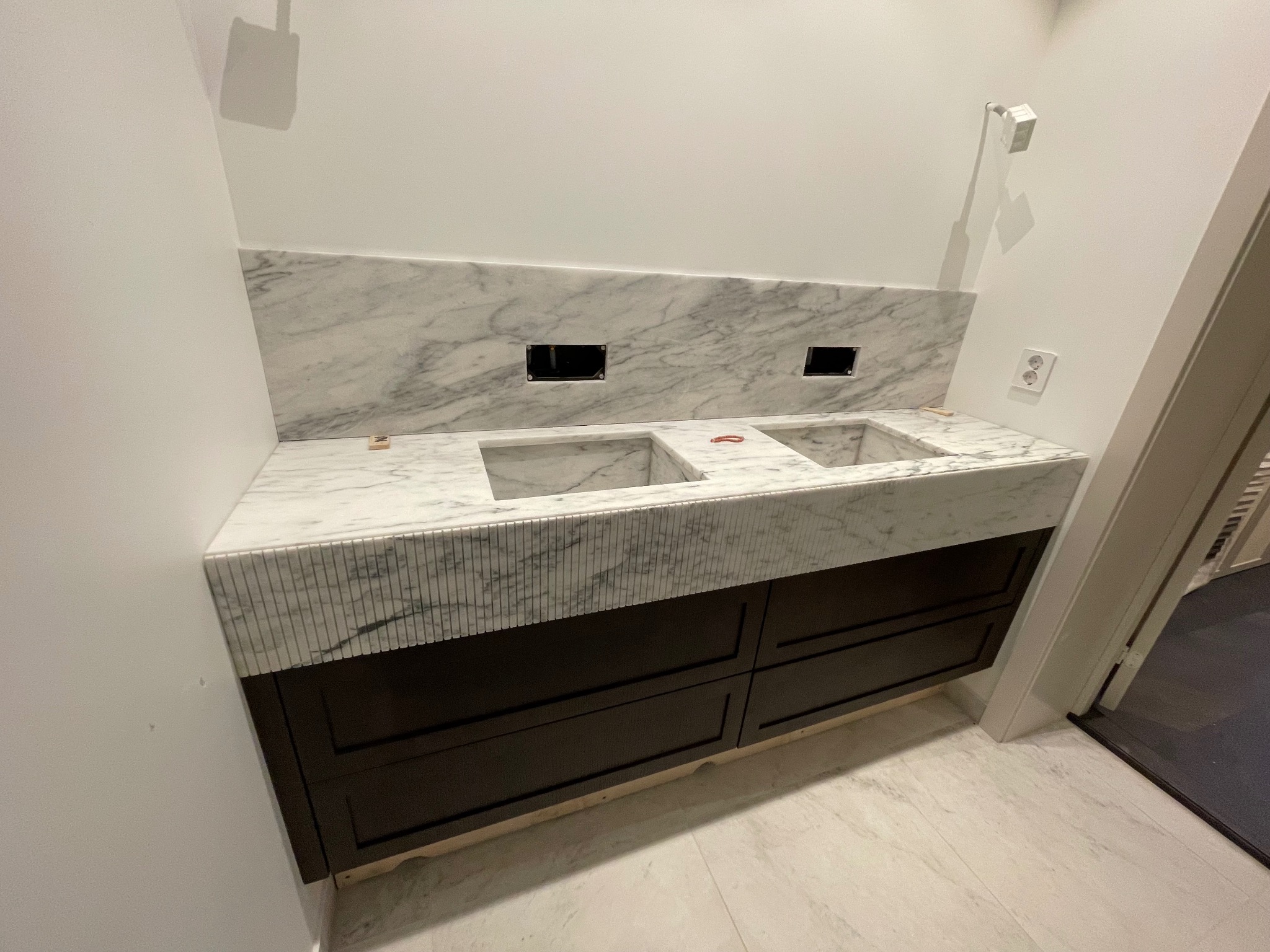 Łazienka w trakcie wykańczania: marmurowa umywalka z dwiema misami i ciemnymi szafkami, widoczne otwory na armaturę, białe ściany i podłoga.