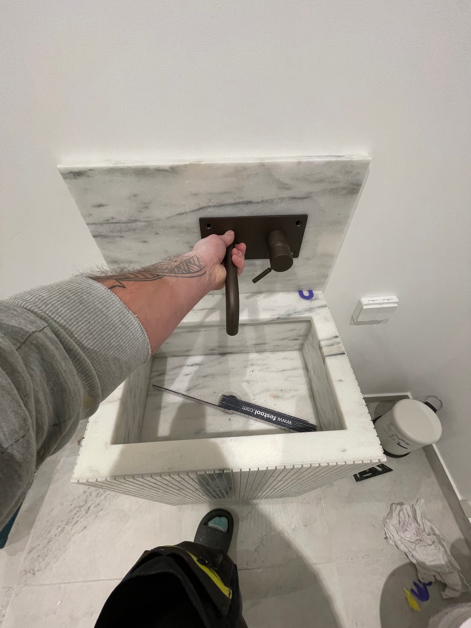 Marmurowa umywalka z brązową armaturą, montowana przez glazurnika. Widoczne narzędzia i fragment podłogi z płytek. Ujęcie z góry, podkreślające detale wykończenia.