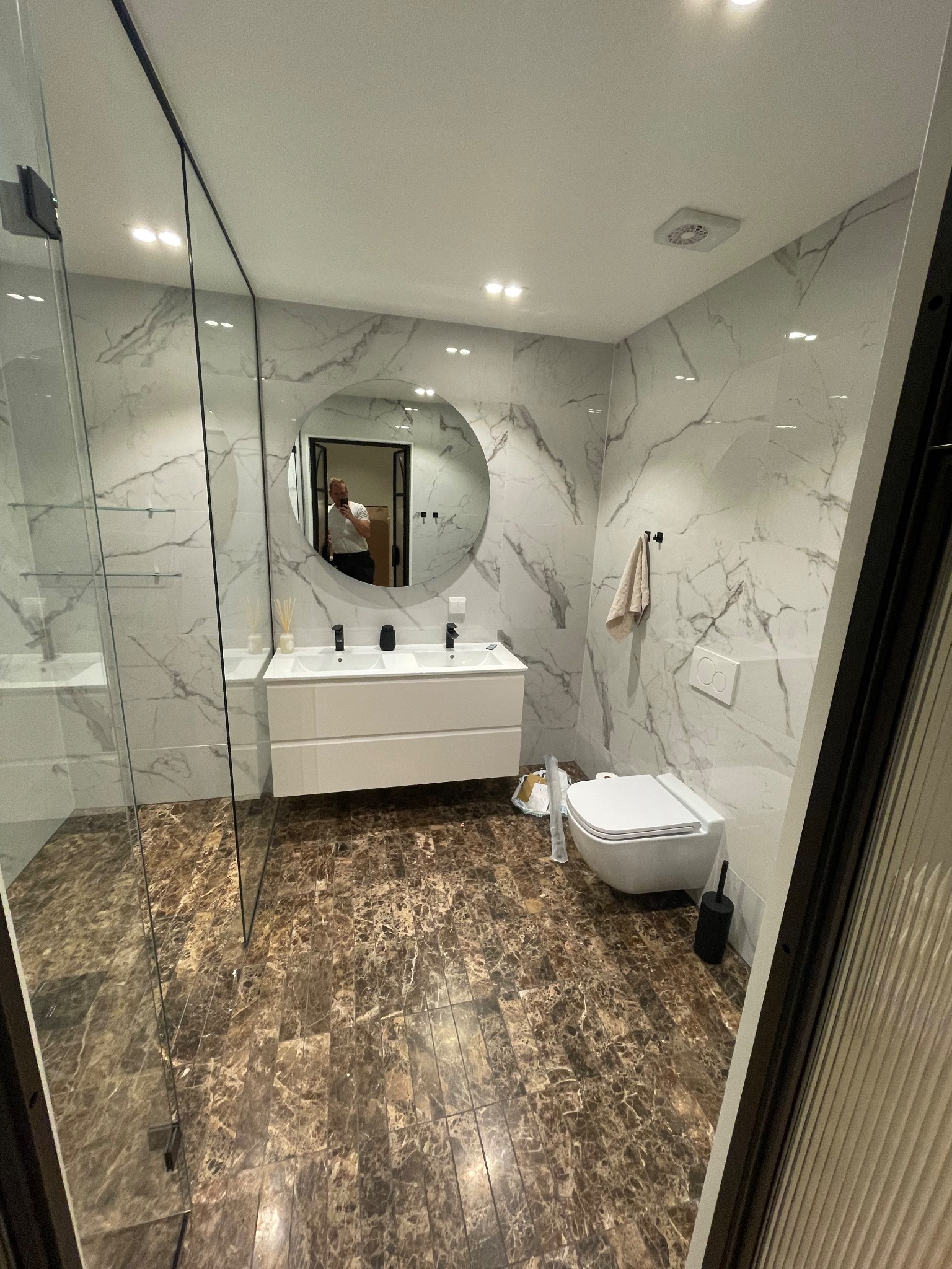 Nowoczesna łazienka z marmurowymi płytkami, brązową podłogą i okrągłym lustrem. Kabina prysznicowa ze szklanymi ścianami, biała umywalka z szafką i wisząca toaleta.