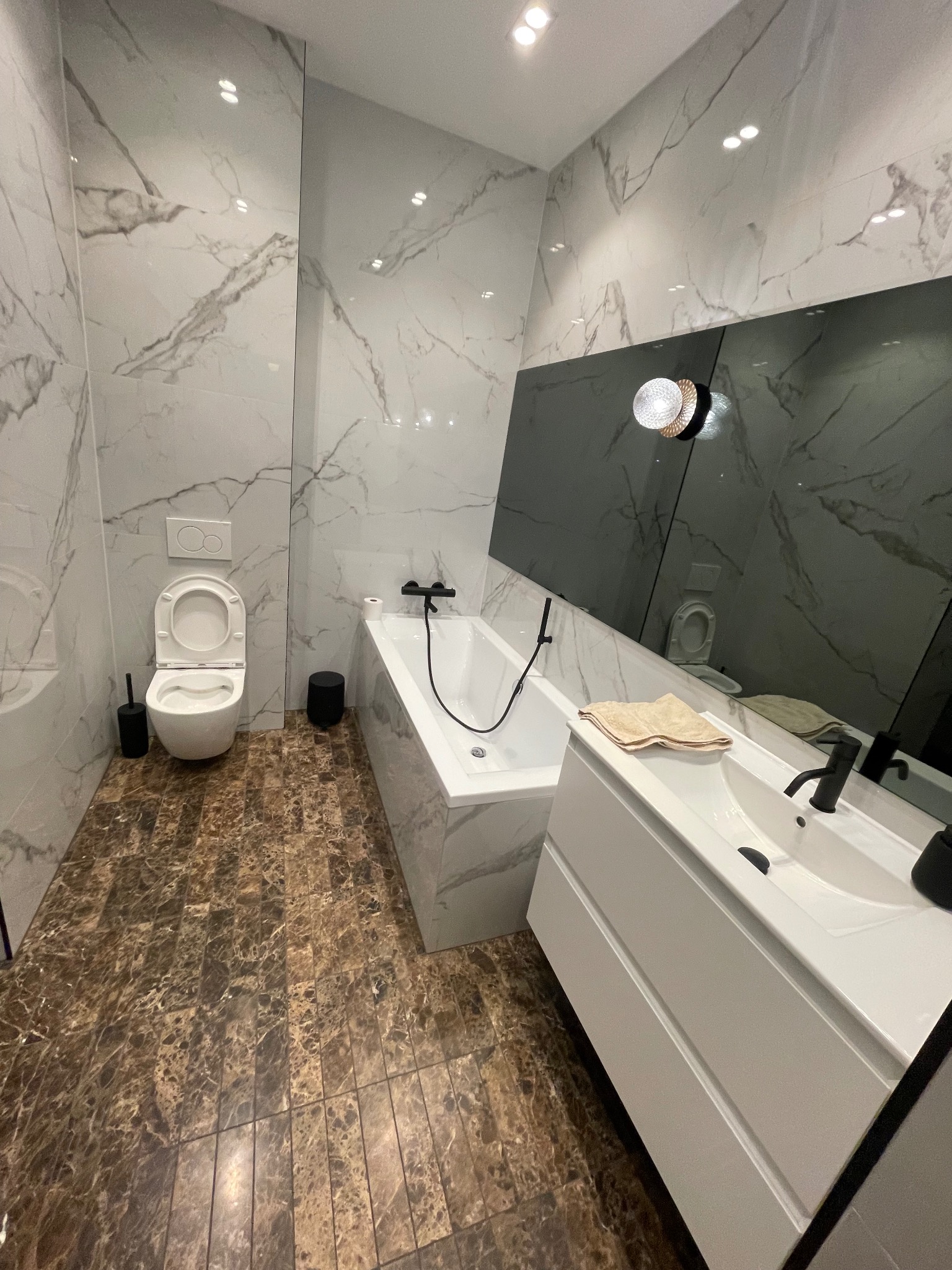 Nowoczesna łazienka z marmurowymi płytkami, brązową podłogą, białą armaturą i asymetrycznym lustrem. Widok na toaletę, wannę i umywalkę z szafką.