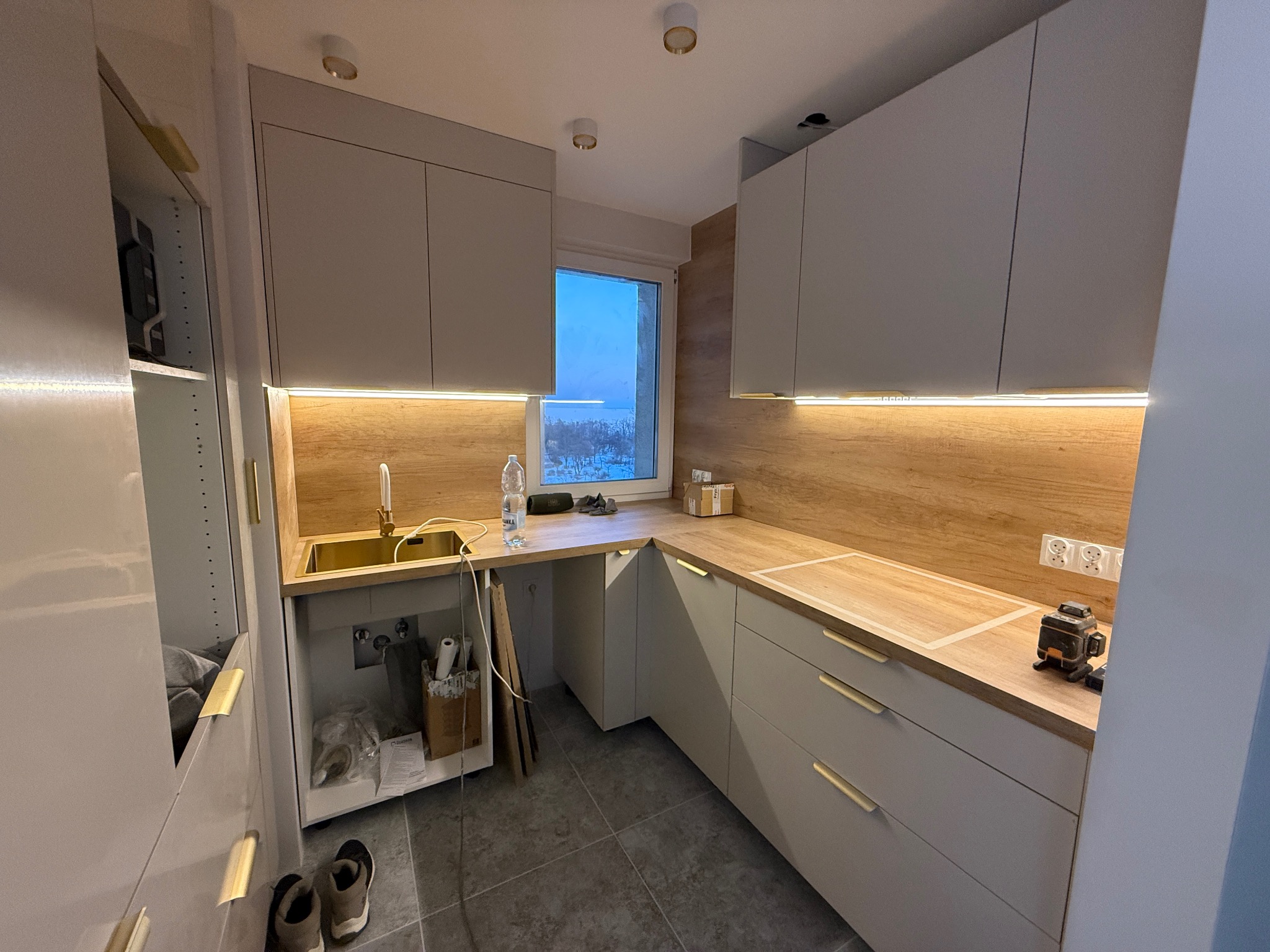 Jasna kuchnia po remoncie z drewnianym blatem i złotymi uchwytami. Widoczny laser krzyżowy na blacie, sugerujący precyzyjny montaż. Nowoczesny design.