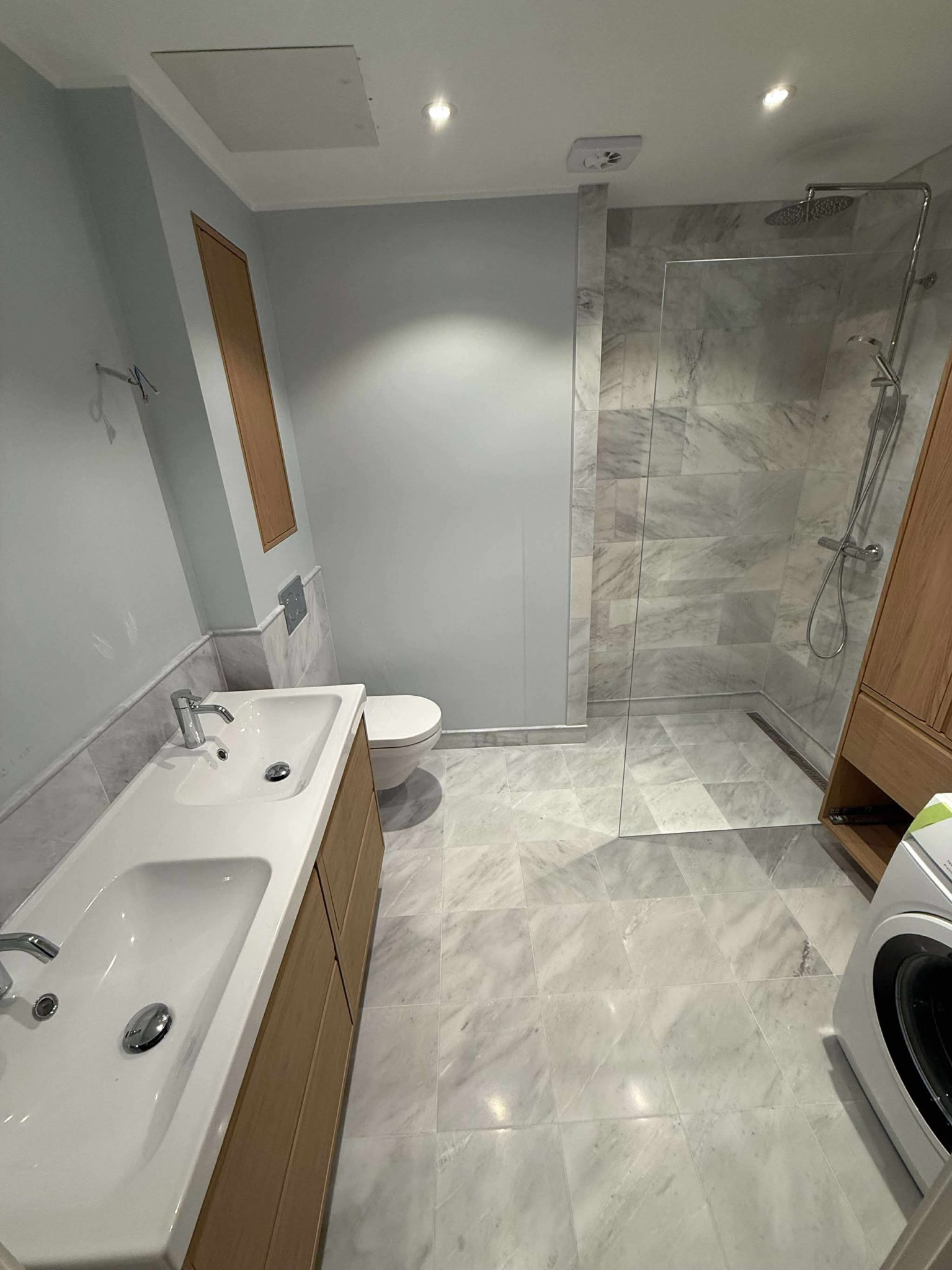 Jasna łazienka z marmurową podłogą i kabiną prysznicową typu walk-in. Podwójna umywalka z drewnianą szafką, toaleta podtynkowa. Minimalistyczny design.