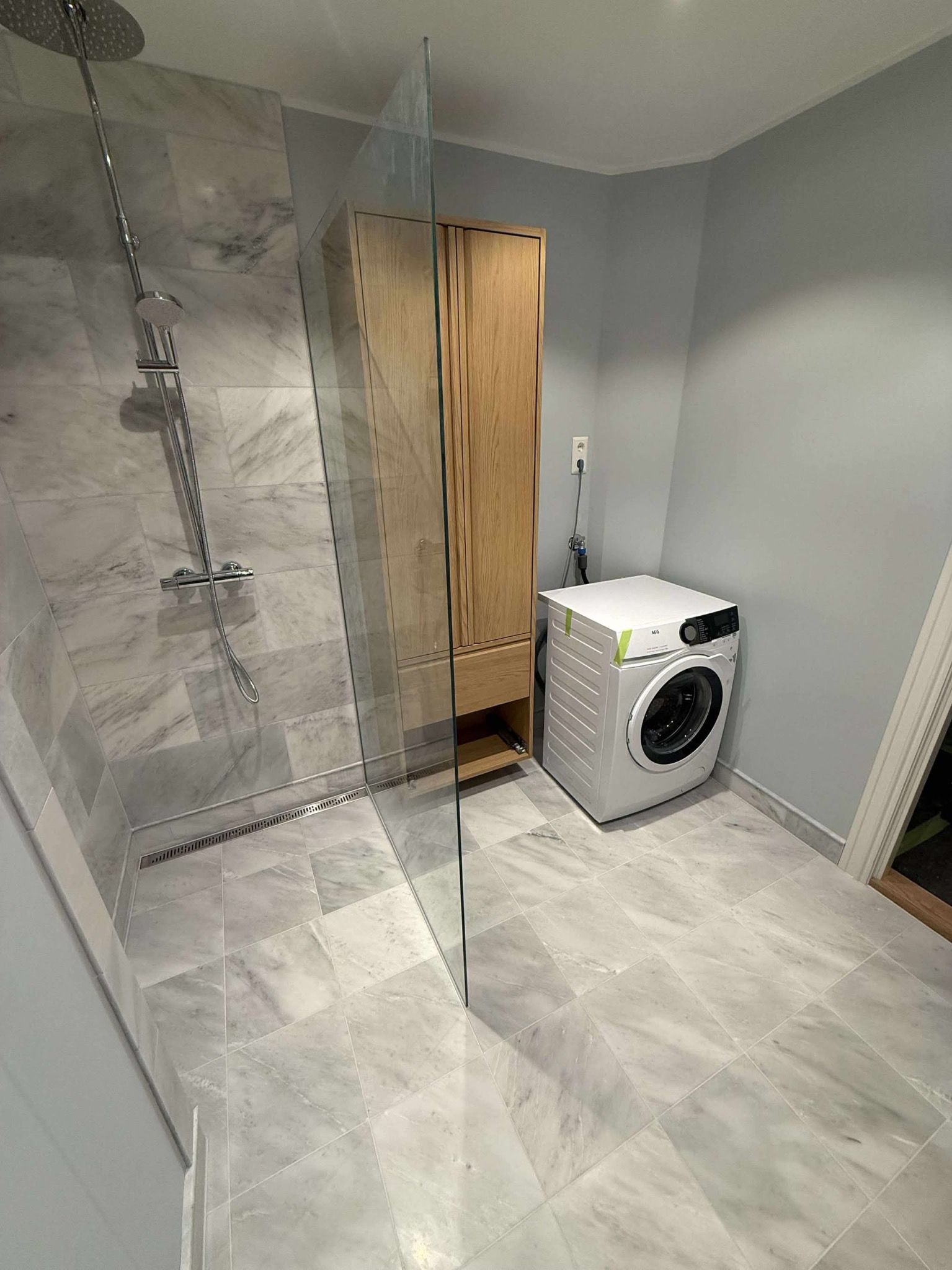 Nowoczesna łazienka z marmurowymi płytkami, kabiną prysznicową typu walk-in, drewnianą szafką i pralką. Szare ściany i podłoga tworzą spójną całość.