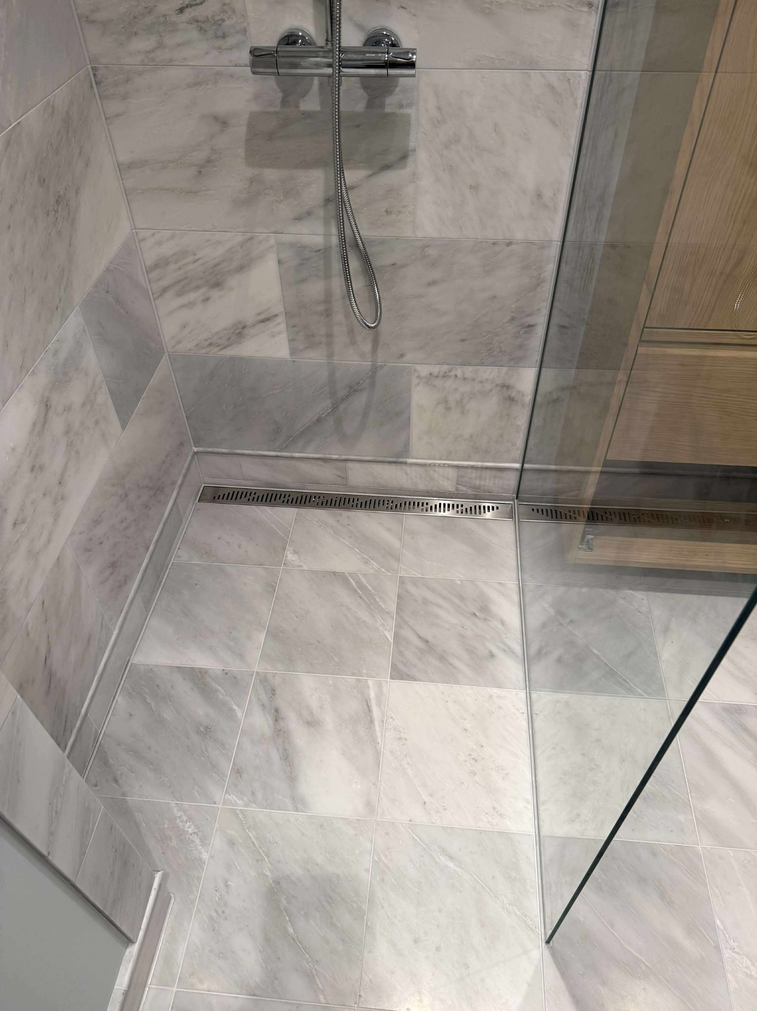 Wykończona kabina prysznicowa z marmurowymi płytkami, chromowaną armaturą i odpływem liniowym. Widoczne szklane drzwi kabiny i fragment drewnianego elementu.