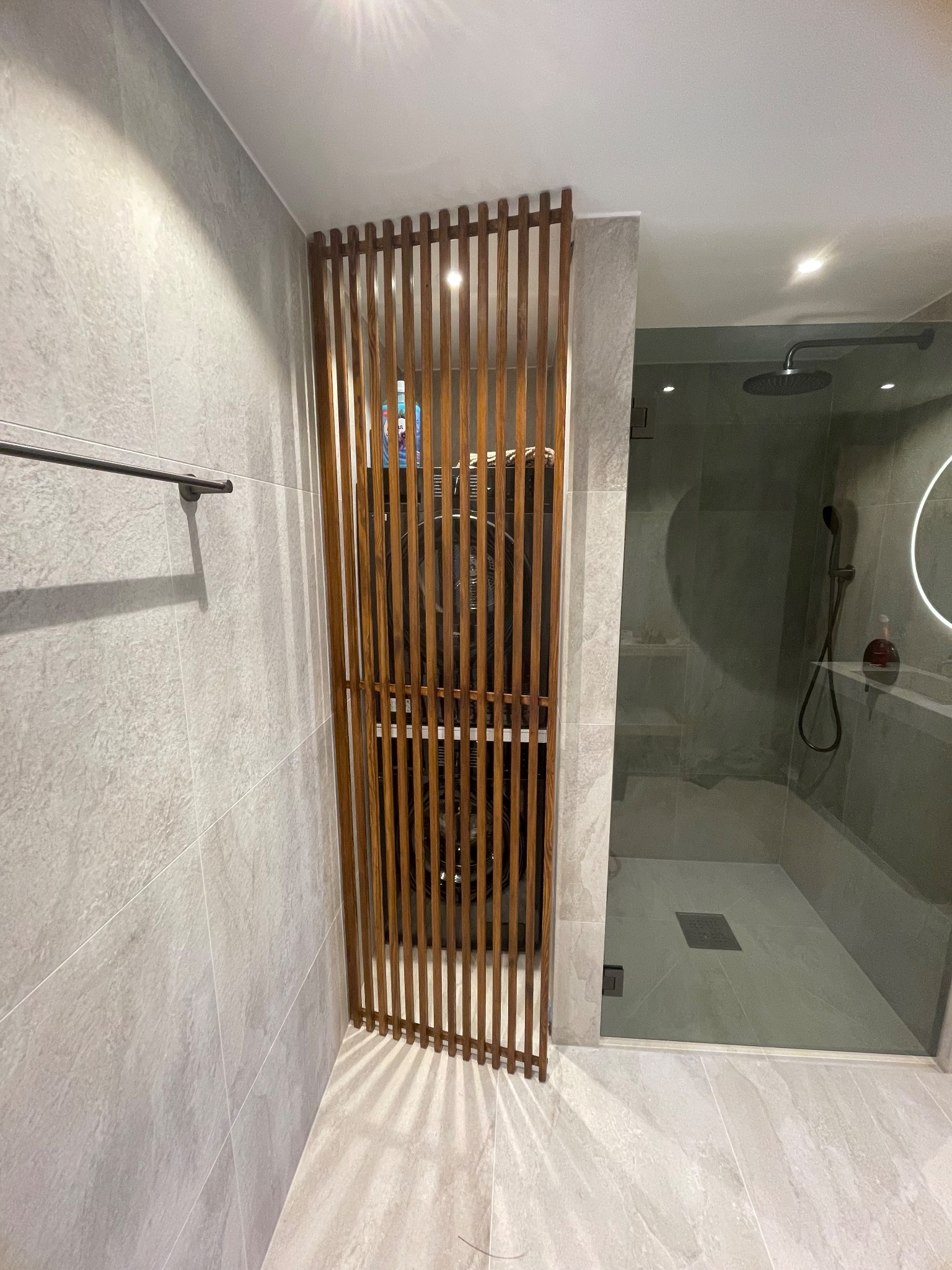 Nowoczesna łazienka z kabiną prysznicową oddzieloną drewnianą ażurową ścianką. Szare płytki na ścianach i podłodze. Minimalistyczny design.