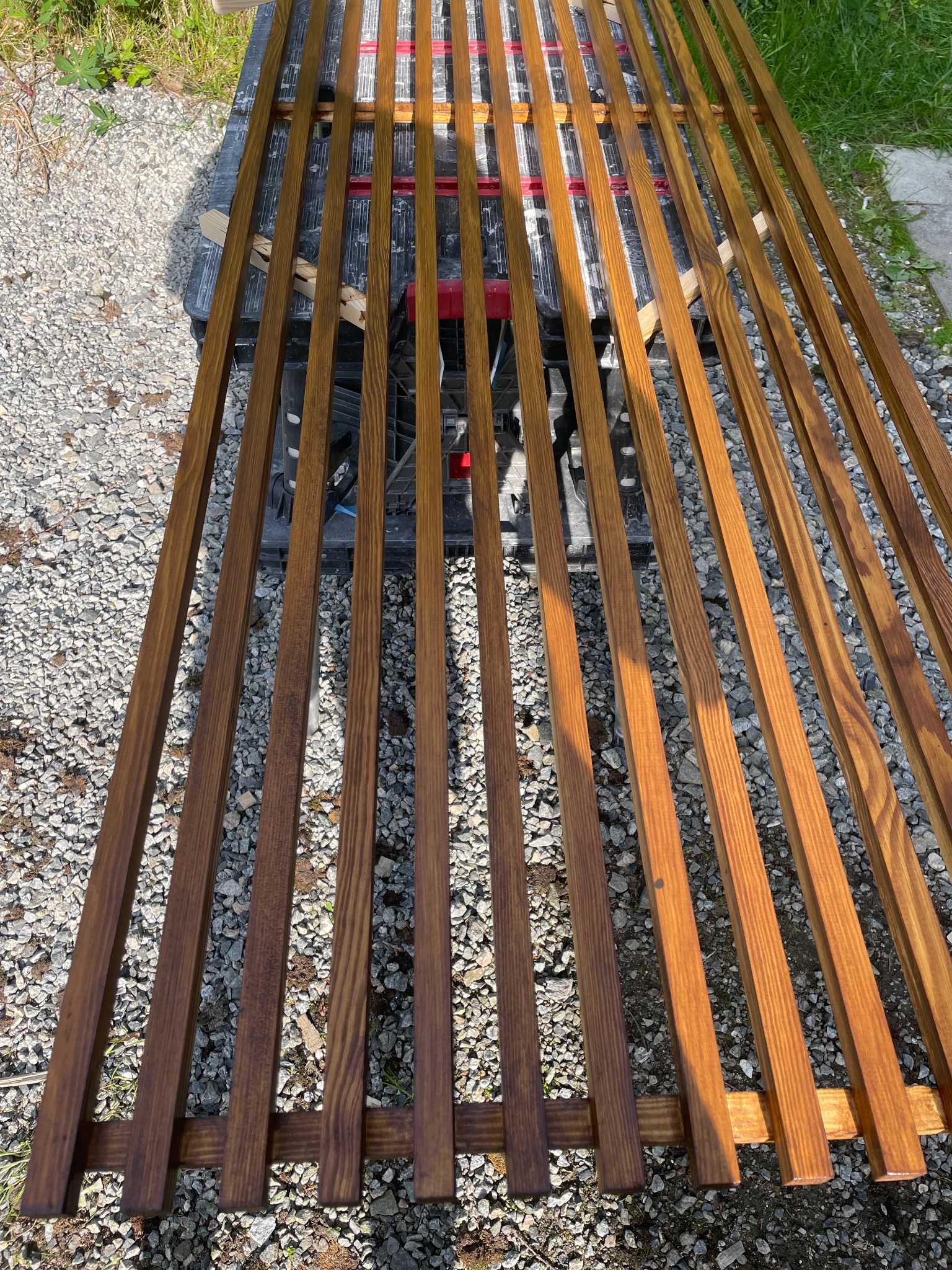 Drewniane, pomalowane listwy ułożone równolegle na czarnym stole roboczym, na tle żwirowego podłoża i trawy. Widoczne słoje drewna i świeży lakier.