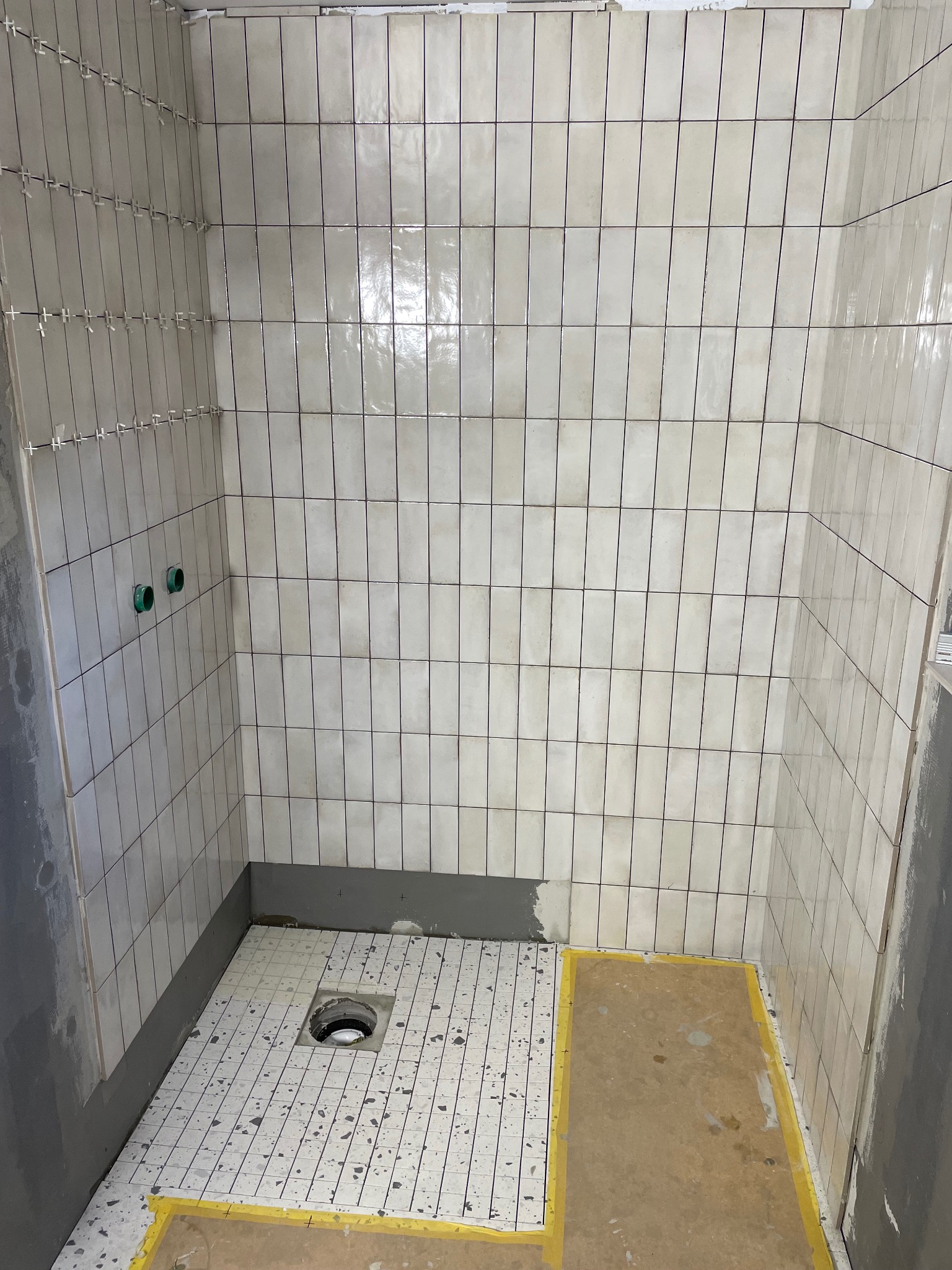 Ściany w łazience wyłożone pionowymi, kremowymi płytkami z krzyżykami dystansowymi. Na podłodze płytki z szarymi plamkami i otwór odpływowy.