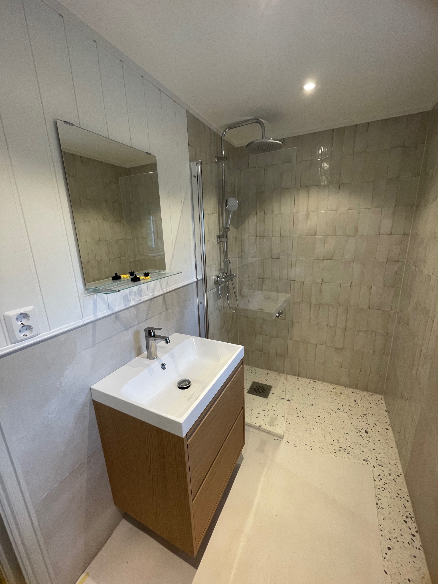 Nowoczesna łazienka z kabiną prysznicową typu walk-in, umywalką z szafką z drewnianym fornirem i lustrem z półką. Ściany wyłożone beżowymi płytkami.