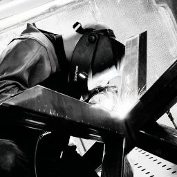 A.I.M Weld Usługi Spawalnicze MARIUSZ PARUZEL - Spawacz w masce ochronnej marki Savage łączy metalowe elementy konstrukcji, iskry rozświetlają kadr. Ujęcie z dołu, czarno-biała tonacja.