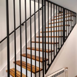A.I.M Weld Usługi Spawalnicze MARIUSZ PARUZEL - Nowoczesne schody z drewnianymi stopniami i czarną, metalową balustradą. Minimalistyczny design, białe ściany, widoczny grzejnik. Ujęcie z dołu, podkreślające geometrię.
