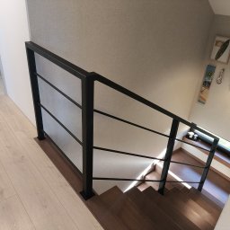 A.I.M Weld Usługi Spawalnicze MARIUSZ PARUZEL - Nowoczesne schody z drewnianymi stopniami i czarną, minimalistyczną balustradą z wypełnieniem z siatki. Widok z góry, jasne wnętrze z dekoracjami na ścianie.