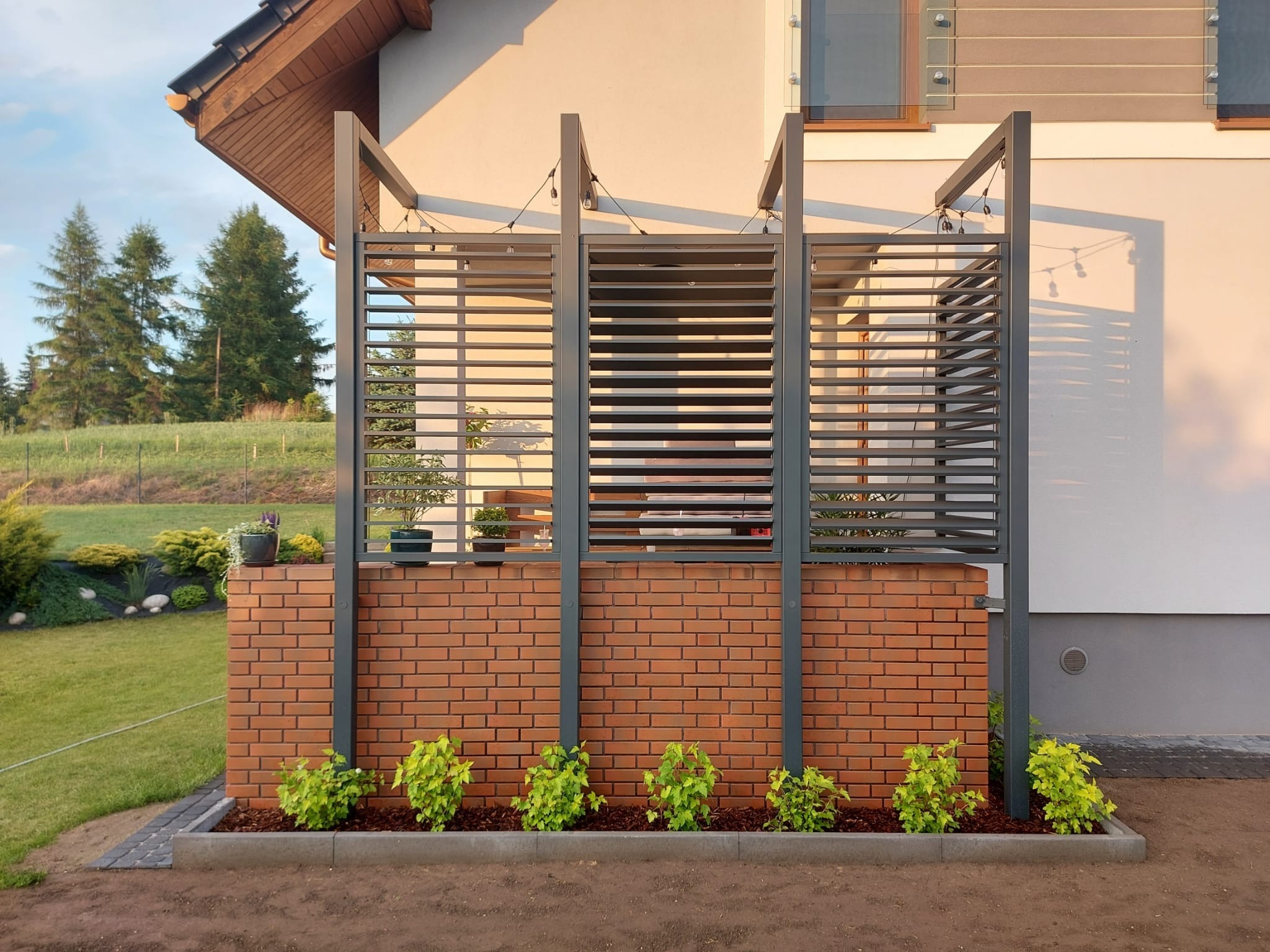 Nowoczesna, szara konstrukcja z poziomych listew, zamontowana na murku z czerwonej cegły, z roślinami u podstawy. Oświetlenie dekoracyjne.