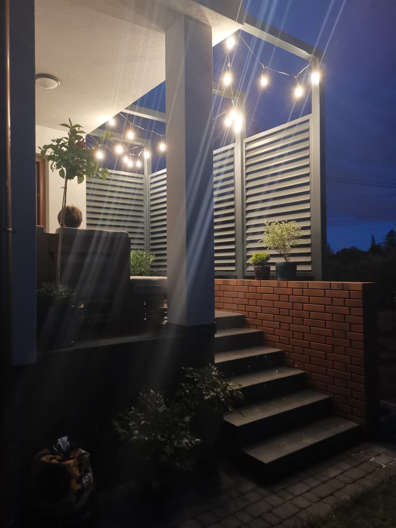 Nowoczesna balustrada z poziomych, szarych profili, oświetlona girlandą żarówek, z widocznymi schodami i roślinami w donicach, tworząca klimatyczny kącik.