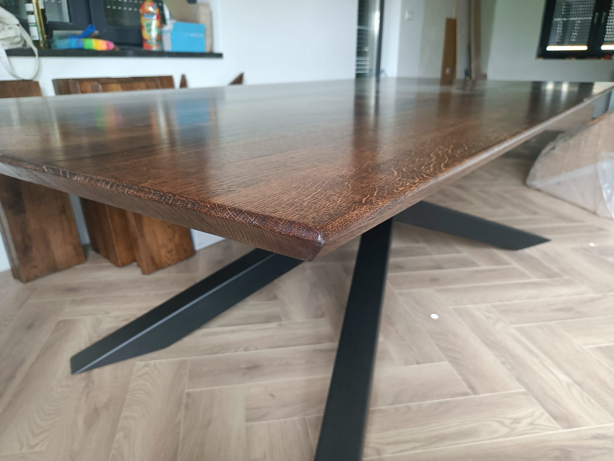 Stół z blatem z ciemnego drewna i czarnymi, metalowymi nogami w kształcie litery X. Widoczna struktura drewna i matowe wykończenie nóg. Ustawiony na podłodze w jodełkę.