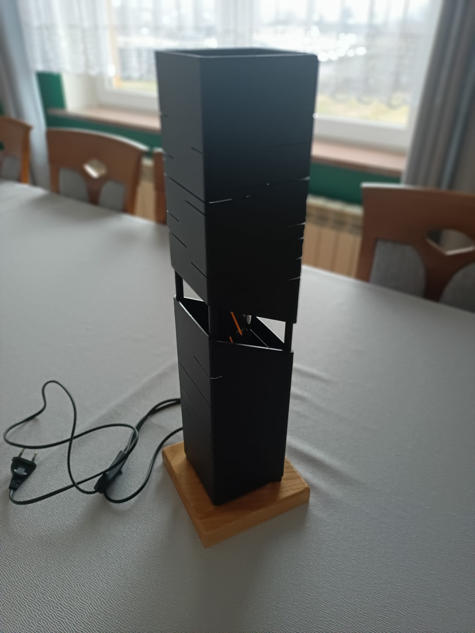 Czarna, nowoczesna lampa metalowa z drewnianą podstawą, stojąca na stole w jasnym wnętrzu. Unikalna konstrukcja z poziomymi szczelinami. Widoczny kabel zasilający.