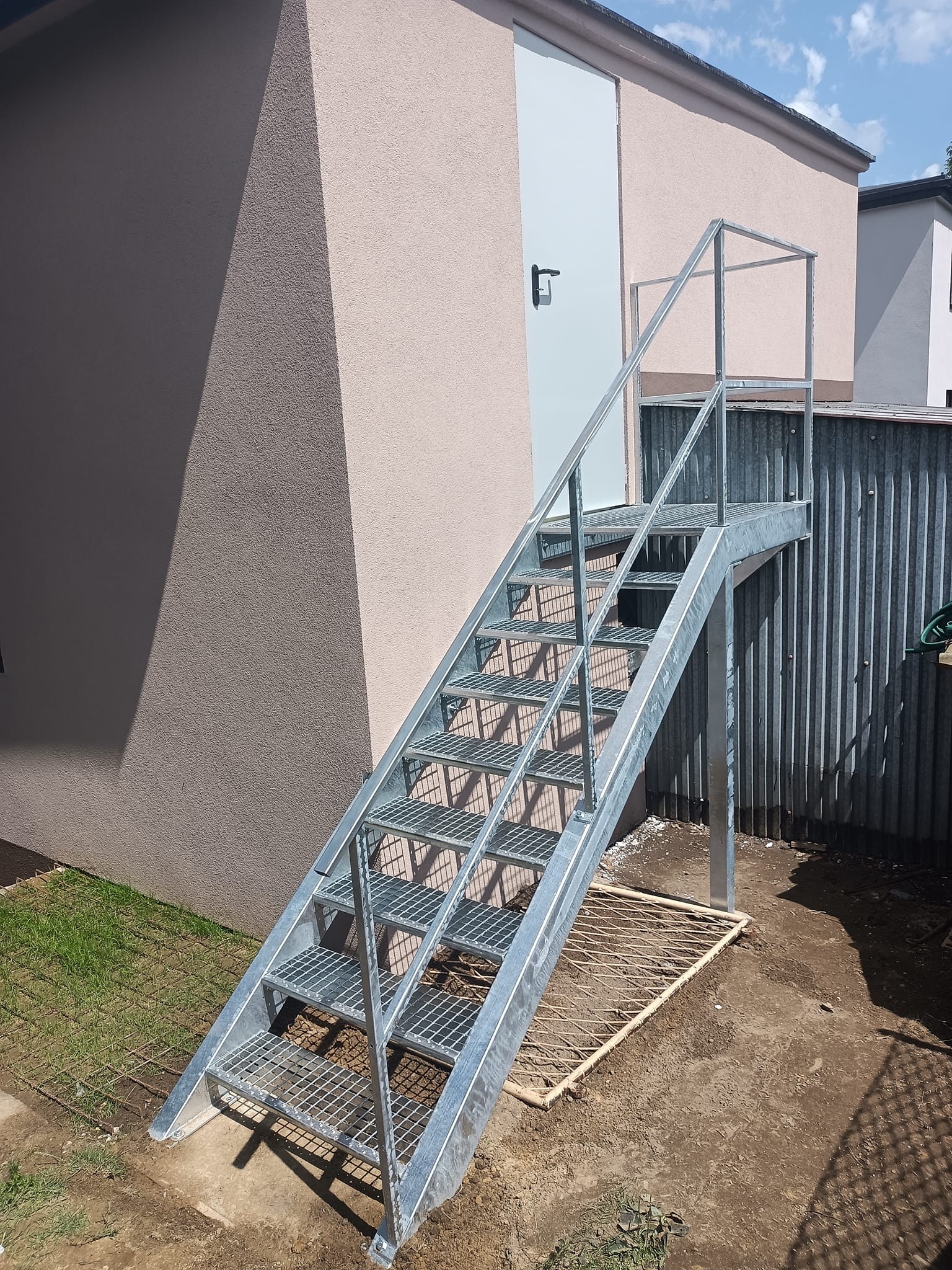 Zewnętrzne schody metalowe z ocynkowaną konstrukcją prowadzące do drzwi budynku. Widoczne kraty stopni i barierka. Tło stanowi elewacja i fragment ogrodzenia.
