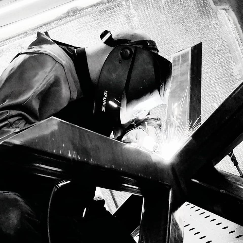 Spawacz w masce ochronnej marki Savage łączy metalowe elementy konstrukcji, iskry rozświetlają kadr. Ujęcie z dołu, czarno-biała tonacja.