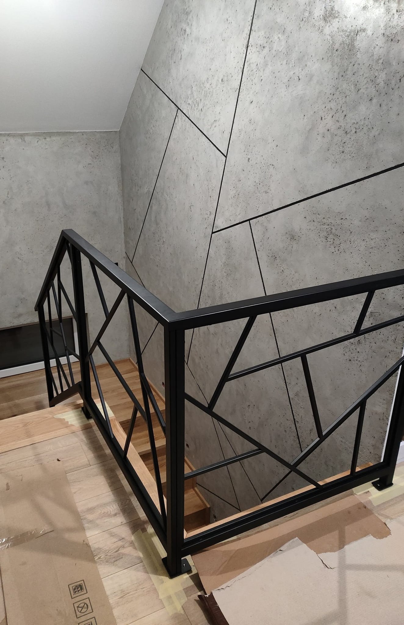 Nowoczesna, czarna balustrada schodowa z geometrycznym wzorem na tle ściany z efektem betonu. Widoczne drewniane stopnie i zabezpieczenia podłogi.