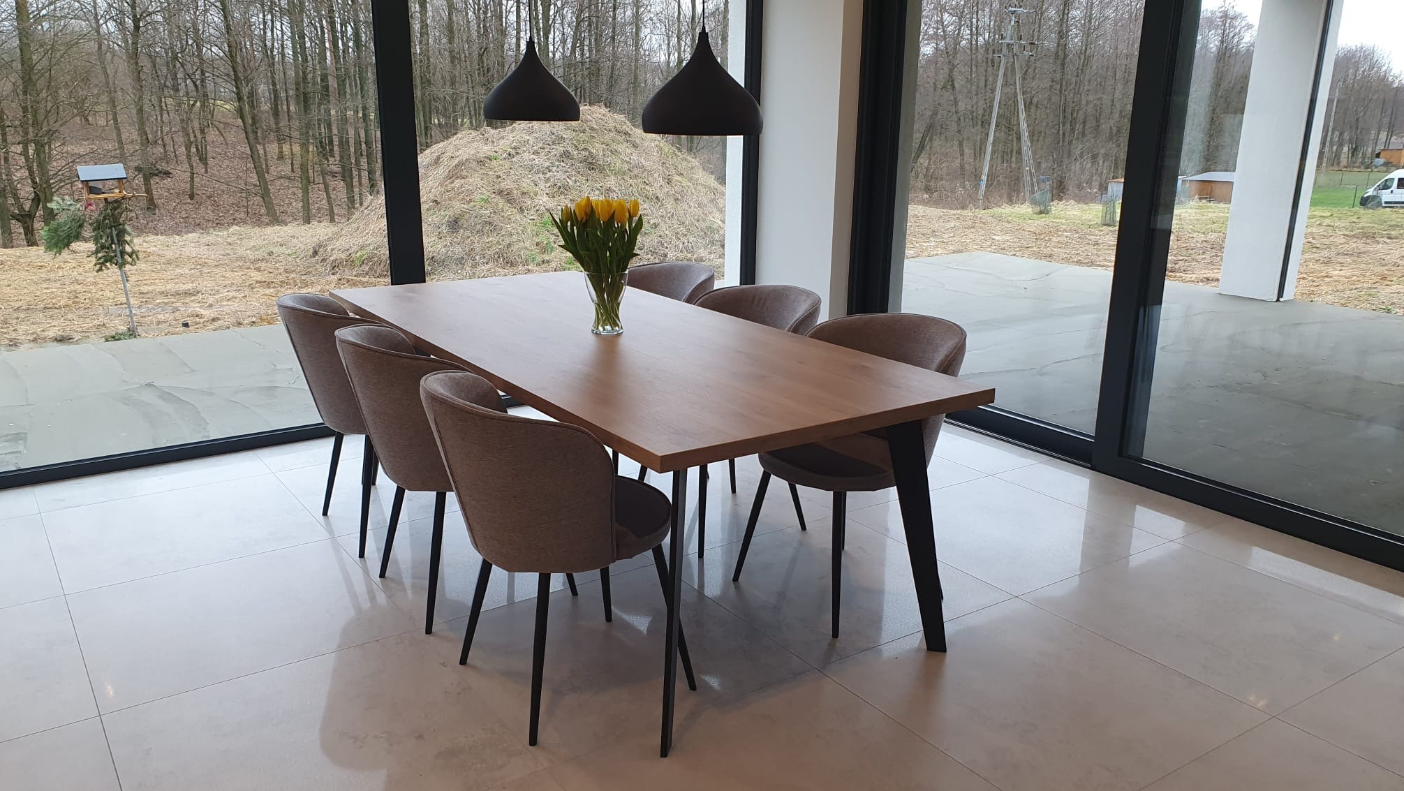 Stół z drewnianym blatem i metalowymi nogami w nowoczesnej jadalni z dużymi oknami. Krzesła tapicerowane w odcieniach szarości. Na stole wazon z żółtymi tulipanami.