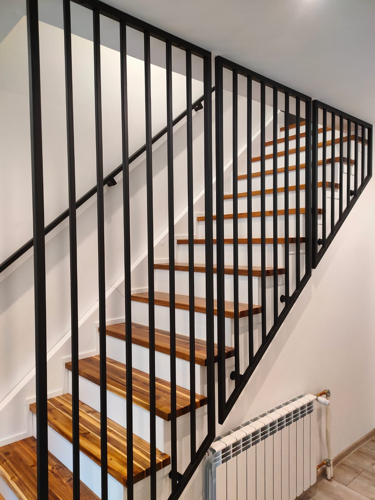 Nowoczesne schody z drewnianymi stopniami i czarną, metalową balustradą. Minimalistyczny design, białe ściany, widoczny grzejnik. Ujęcie z dołu, podkreślające geometrię.