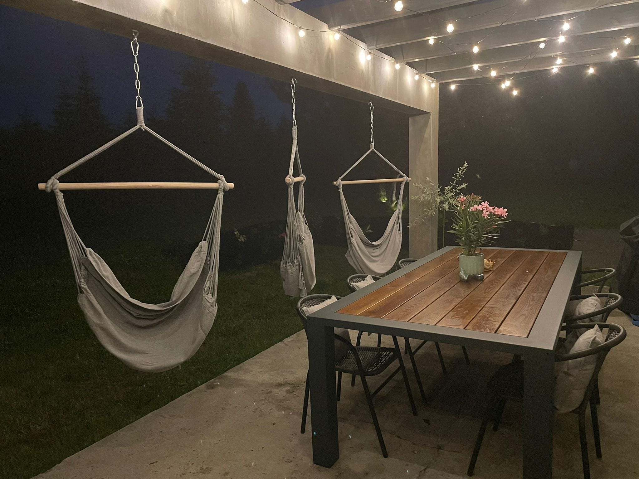 Zadaszony taras z trzema wiszącymi fotelami hamakowymi na łańcuchach, oświetlony girlandą żarówek. Stół z kwiatami i krzesłami. Wieczorny klimat.
