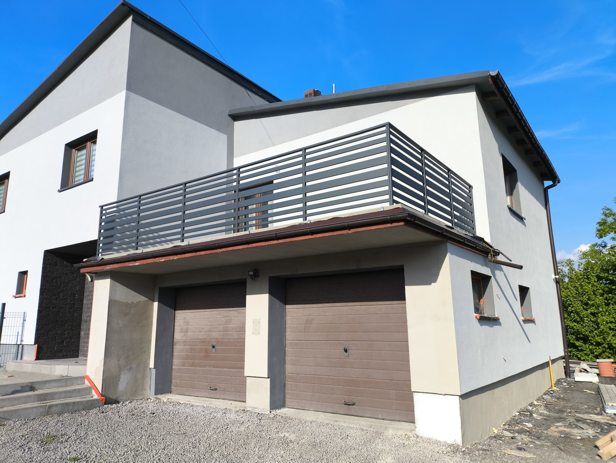 Nowoczesna balustrada balkonowa z poziomych profili na tle dwukondygnacyjnego domu z garażem i błękitnym niebem. Minimalistyczny design, jasna kolorystyka.